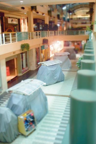 No. 4, “a-Hunting.” The second-floor balcony of an empty shopping mall, at night; the lights are low, and the storefronts dark, except for one down on the ground floor, midway down the length of the mall, shining too brightly to make out anything within. Past it, a bridge links the balconies on either side beneath arched glass ceilings. The kiosks out on the floor below are draped with pale blue cloths, waiting for tomorrow.