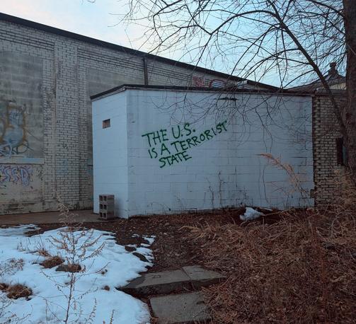 Photo of a small cinderblock outbuilding tagged with the sentence, "THE U.S. IS A TERRORIST STATE" in green spray paint.