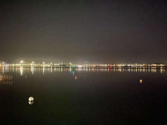 Nächtliche Förde mit durch weiße Bojen eingegrenzter Badezone, einer nahen grünen Tonne und entfernten roten Tonne die als Schifffahrtszeichen fungieren und dem hell erleuchteten Marinearsenal am Kieler Ostufer, welches als Lichtband das Bild in der Mitte von links nach rechts durchzieht. Förde und Himmel erscheinen im Kontrast dunkel, in den Ecken des Bildes fast schwarz. Die Lichter am gegenüberliegenden Fördeufer sind einheitlich gelb, nur die zwei Tonnen stechen mit ihrem rot und grün als Farbtupfer hervor.