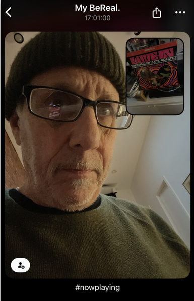 ChatGPT sez:

This screenshot appears to be from the BeReal app. [It is.] It shows a selfie of a man wearing glasses, a dark knit cap, and a dark green sweater. [It’s me!] The background includes part of a room with white walls and a visible doorway. The lighting is warm and slightly dim.

In the top right corner of the image, there’s a smaller photo (the secondary BeReal image), showing a vinyl record on a turntable. The record is “Love-In” by The Charles Lloyd Quartet, with its colorful, psychedelic-style album cover visible.

At the top of the screenshot, it says:
	•	“My BeReal.”
	•	“17:01:00” (indicating the time the BeReal was posted).

At the bottom, there’s a hashtag:
	•	#nowplaying, likely referencing the album being played.