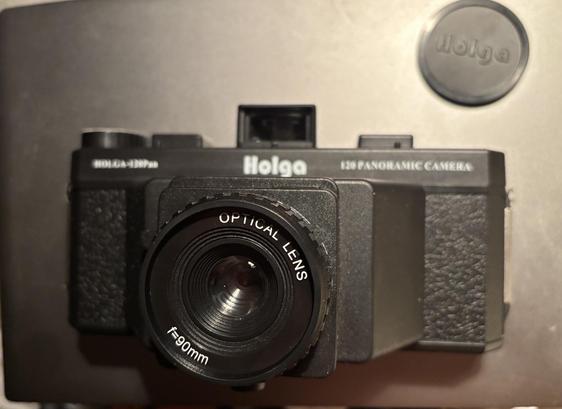 The front of a medium format plastic lens camera, but it’s a panoramic format. The plastic lens is seen, as is the lens cap with brand name “Holga”.