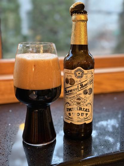 A stout glass half full of beer and half full of thick tan foam. The brown bottle has a gold foil neck wrap, with a white label with black and shiny gold lettering.