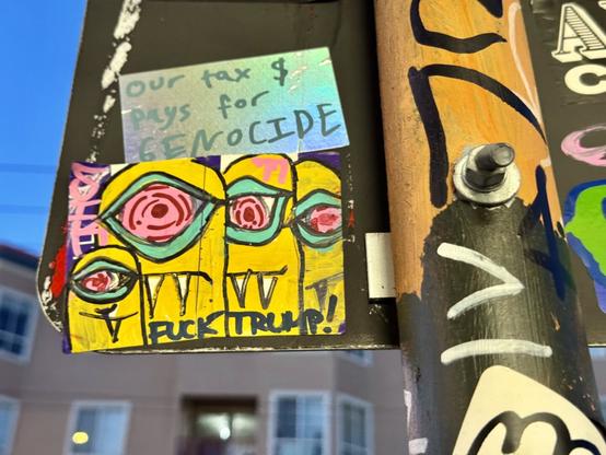 Stickers on a pole on a busy street. One of the stickers has four yellow monsters that sort of look like evil Minions. They have red cyclops eyes and sharp fangs. The name "Sik" is written on the left side and "Fuck Trump" is written on the bottom. There are other hand-drawn sticker slaps around this sticker.