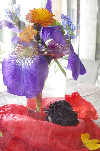 A shot glass sits on a large red poppy leaf in a baseless glass cylinder.
The almost black spore capsule including stamens of the same colour and a yellow buttercup flower were placed in front of the glass.
The glass contains a blue-purple iris flower, a purple closed thistle flower, three buds and two flowers of orange hawkweed and speedwell tendrils. Part of the water surface appears red because it reflects the poppy leaf. The background looks as if it was painted because of the cylinder; It is unobtrusive white, beige and gray, the bright colours dominate it.