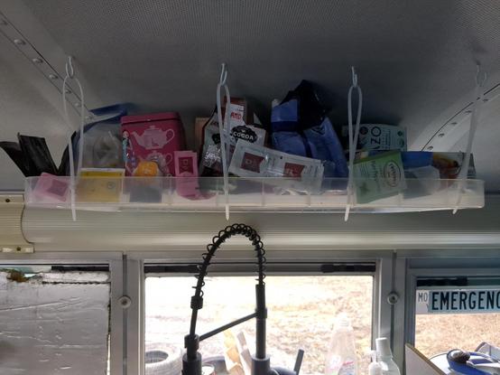 A shelf over a sink fitted inside a bus, the shelf is made of a plastic box lid hanging from cable ties on hooks in the bus ceiling. The shelf is full of boxes of teabags amd associated items such as small sachets of hot chocolate and teabags that escaped their boxes. The window to the right of the sink is marked ad an emergency exit. The window to the left has a slightly damaged piece of old insulation board roughly positioned over it.
