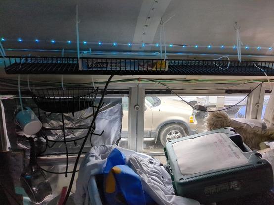 A wire shelf hung from the ceiling of a bus using cable ties, with a roll of aluminium foil in a dispenser box on the shelf. A wire mug tree is hung in a similar manner against the window below the shelf to its left side. A mug and saucepan are already hanfing from that. A green case for a power drill is closed and placed atop other stuff in the foreground, marked with whose property it is (a list of several names).
Vehicles can be seen out of the window.
A string of blue LEDs is stuck to the ceiling above and behind the shelf.
The window behind the mug tray is covered with foil. The window to the right has glue deposits from duct tape that failed to hold a rug in place as insulation over the winter.