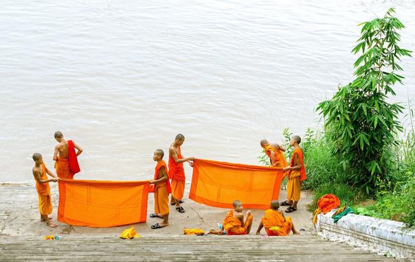 Photo shows a group of young buddhist monks/novices doing laundry of the deep yellow-orange robes typical for them. They are standing on a platform with the Mekong river flowing greyish in the background, and the photo is taken down the stairs to that platform. One of the youngest member of the group has turned his head and is looking straight at the photographer. Some small, green trees and bushes are visible to the right in the photo.