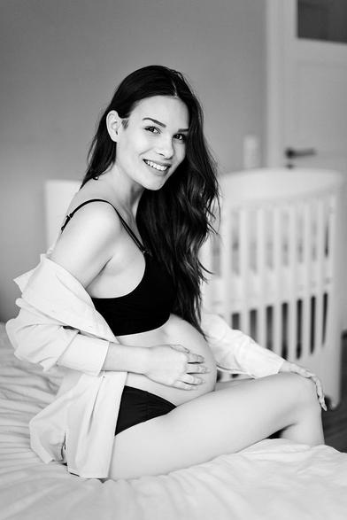Pregnant woman in underwear sits on her bed and smiles into the camera