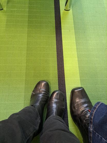Schuhe an Füßen auf einem grüngestreiften Teppichboden - typisches Wartezimmerfoto.