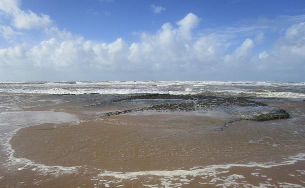 Rivage avec un rocher rond et plat qui émerge des vagues et du sable. Ciel bleu nuageux