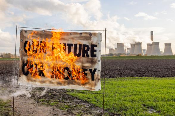 Outdoors in a field, banner 'Our Future our money' is on fire, with active cooling towers of wood-burning power station in the distance