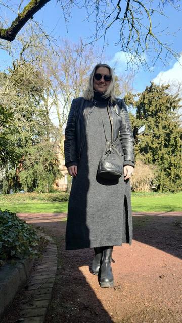 ENG A woman standing in the sun, wearing a long dark grey knitted dress, a black biker jacket and black boots.

DEU Eine Frau steht in der Sonne und trägt ein langes dunkelgraues Strickkleid, eine schwarze Lederjacke und schwarze Boots.
