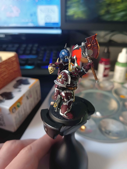Campeón marine espacial del Caos del juego de mesa Warhammer 40k pintado en el esquema de color de los Portadores de la Palabra (burdeos con detalles en plata, aunque yo he añadido algunos detallitos en dorado también). Del casco le salen cuernos pintados en plata y hueso, y de los extremos de su mochila salen dos calaveras pintadas en blanco hueso. Sobre esa mochila clavada en un pincho hay un casco azul (del capítulo de los Ultramarines) con una calavera en su interior. En su mano derecha sujeta una pistola bólter con la estrella del Caos grabada en oro cerca del cañón, y con su mano izquierda alza un hacha cuya hoja está pintada de rojo oscuro con colores más claros en el filo para dar la sensación de que está en llamas.