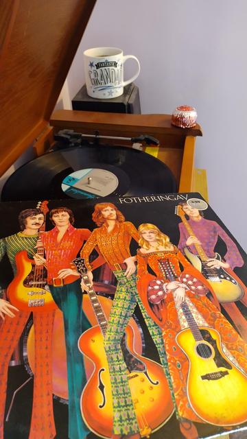 Record player with a 12" black vinyl record.
In the foreground is an album cover featuring a drawing of the five band members in brightly coloured clothes and holding guitars.
Behind the player is a mug of coffee on a speaker. On the corner of the player is a delicious Tunnock's Teacake.