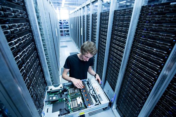 A technician is seen in a hallway of rack servers. He is working on a server that has been pulled out of the nearby rack