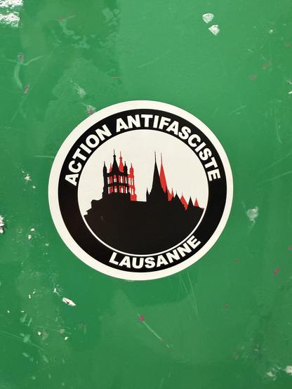 Lausanne’s cathedral, known for its contribution to the city’s skyline, drawn in the typical antifa black and red variation, in a circle, with the french-speaking mention « Action Antifasciste Lausanne ». On a green wall. Lausanne, Vaud, Switzerland, Passage sous-rue St-François, février 2025