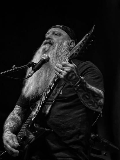 This is a black and white photograph of Kirk Windstein, from the band Crowbar, a heavily tattooed musician with a long, flowing beard, playing an electric guitar on stage. He is wearing a backward baseball cap and a dark T-shirt. His facial expression conveys deep focus and emotion as he sings into a microphone positioned in front of him. His left hand is pressing the frets on the guitar neck, while his right hand is strumming the strings. The guitar has an angular body shape, likely suited for rock or metal music. The background is dark, emphasizing the musician as the focal point of the image.