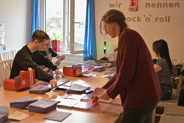 Auf dem Bild sind vier Personen zu sehen, zwei männlich gelesene und zwei weiblich gelesene. Die vier Personen stehen und sitzen jeweils um einen großen Tisch herum, auf dem mehrere Umschläge und DIN A5 große Karten ausliegen. Einige der Personen greifen nach Karten, während andere die Karten in Umschläge stecken.