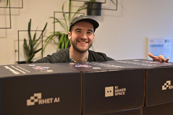 Auf dem Bild ist eine junge, männlich gelesene Person zu sehen, die in die Kamera lächelt. Vor der Person stehen drei schwarze Boxen, auf die je abwechselnd einmal das Logo des RHET AI Centers und des KI Makerspace gedruckt sind.