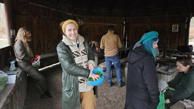 Smiling person in many layers of warm clothes holds a blue bowl of green leaves and is massaging them. Other people are active in the background. Inside a wooden hut in the woods.