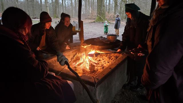 Many people sit around a fire with snobrød on sticks. Woods and children playing in the background.