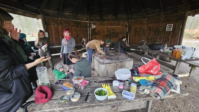 Many people in wooden fireplace shelter in the woods. An adult and a child start a fire in the pit. Someone licks their fingers. Someone massages leaves in a plastic bag. Foods and gloves on a picnic table.