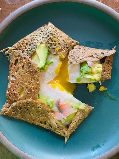 a delicious lacy buckwheat crepe with a fried egg inside topped with avocado and smoked salmon. it's resting on a blue ceramic plate and the yolk is broken.
