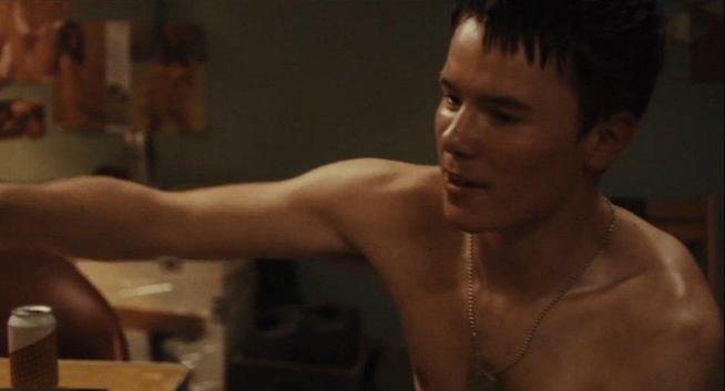 Edvin Ryding dans une scène de caserne de A life's Worth. Torse nu, relativement musclé, en sueur, collier avec plaque militaire, cheveux sombres (mal) coupés courts. Canette de bière sur la table, posters de filles sexy (flous) dans le décor.