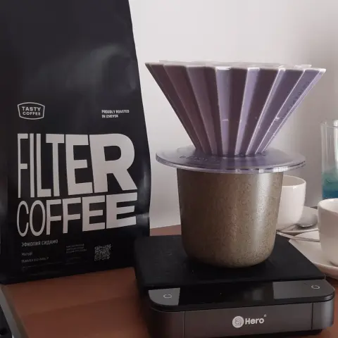 The image showcases a coffee brewing setup featuring a purple plastic coffee dripper with a ridged design sitting atop a speckled ceramic mug. The dripper is placed on a transparent plastic base. This arrangement rests on a black digital scale branded "Hero," which is positioned on a wooden surface.  

To the left of the brewing setup, there is a black bag of coffee labeled "FILTER COFFEE" in bold white letters. The bag also includes additional text in Russian, suggesting details about the coffee’s origin and roasting process.  

In the background, several white ceramic cups and a glass are visible. The scene is set against a plain white wall, and the lighting is soft and natural. The composition highlights the coffee-making process, with a clean and minimalistic aesthetic.
