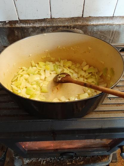 Sur un poêle à bois, une cocotte en fonte noire contenant des oignons en train de revenir tranquillement. Une cuillère en bois sur le bord.