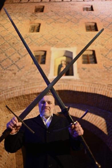 Zdjecie Adriana Zandberga z dwoma długimi skrzyżowanymi mieczami w dłoniach. W tle ściana budynku z cegły.