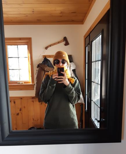 Mirror selfie of the artist. Her face hidden by big round shades, a yellow scarf and her phone