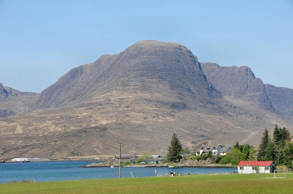 The mountains of the Applecross Peninsula. The image shows a green field in the foreground leading back to a blue loch with some buildings on the right if it. Beyond the loch is a range of dark rocky mountains of considerable height. The sky is blue.