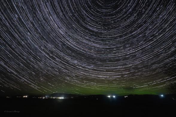 A night sky time lapse image showing a black foreground with scattered lights from houses  and low hills. The sky above the horizon is glowing green to varied degrees but there are white streaks  like the grooves on a vinyl record caused by the rotation of the Earth. Several hundred photographs all stacked together to give this effect,