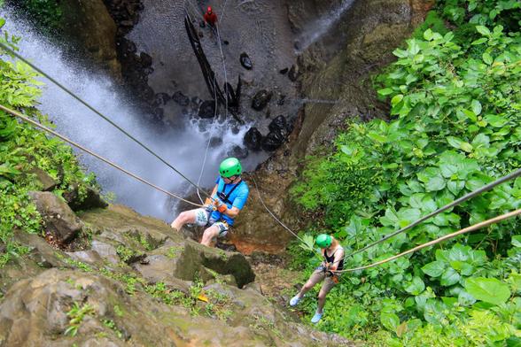 the view is from the top looking straight down a deep waterfall. two people are on ropes half-way down, wearing safety gear, rappelling down to the bottom, where a guide awaits them. the misty spraying waterfall is on the left, lush green leaves are on the right.
