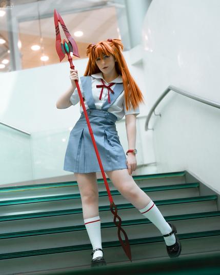 A cosplayer dressed as Asuka Langley Soryu from Neon Genesis Evangelion poses on a modern glass staircase. She wears Asuka’s signature blue and white school uniform with a red ribbon, along with long orange hair styled into twin tails. She holds a red Spear of Cassius prop, standing with a confident stance. The setting is an indoor area with sleek white walls and glass railings.

tag ignore: cosplay cosplaygirl girl kofi throne wishlist request commission
