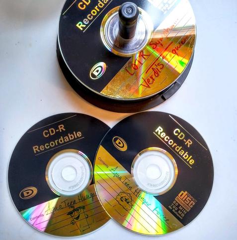 A stack of CD-R discs, shiny gold and black, seen from above on a white surface. The one on the top of the stack is labelled LOTR Symphony and Verdi’s Requiem. Two other discs lay overlapped beside it. They are both labelled Aqua Teen Hunger Force with a drawing of Shake and Meatwad, respectively.