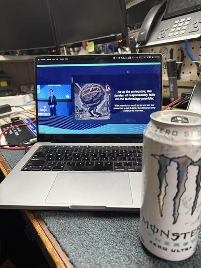 The image shows a laptop on a desk displaying a presentation related to compliance. There is also a can of Monster Energy Zero Ultra on the desk. The setting appears to be a workspace with various equipment and tools visible in the background.