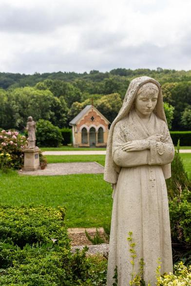 Au premier plan, une statue représente une jeune fille, menton baissé, foulard sur la tête, en habit catholique.
Notre-Dame de Garaison - le jardin du sanctuaire, 2023.