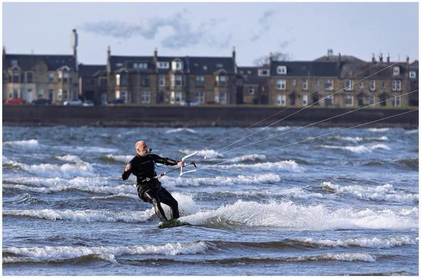 A person is kitesurfing on a wavy body of water, wearing a wetsuit. In the background, there are buildings along the shore under a partly cloudy sky. He has a GoPro camera in his mouth.
