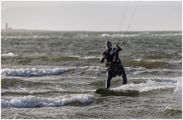 A person kitesurfing on choppy water, wearing a wetsuit and harness, with a kite in the sky above. The background features a distant lighthouse and cloudy sky.