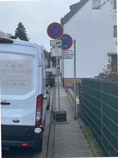 Blick auf einen sehr schmalen Bürgersteig, welcher rechts durch einen Metallzaun, im weiteren Verlauf Mauer und Häuser, und links durch parkende Autos auf einer Parkfläche begrenzt wird. Die Breite des Hochbords beträgt etwa 80-85cm. Es ist ein Mast mit VZ 286-30 (Eingeschränktes Haltverbot mit Pfeilen in beide Richtungen) und dem Zusatzschild "Parken in gekennzeichneten Flächen erlaubt" fest montiert. Auf dem Fußweg wurde zusätzlich ein Mobiles Schild VZ283 (absolutes Haltverbot) mit dem IMO redundanten Zusatz VZ1030-31 (Haltverbot auch auf dem Seitenstreifen) und einem laminierten und sehr verknitterten Papier "Sonntag 2. März 11:00-13:00"(?). Durch die Fußplatten des Schildes verengt sich der Fußweg auf ca. 25cm.
