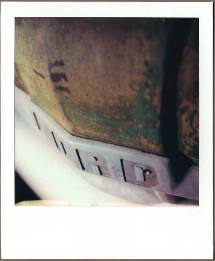 Close-up Polaroid of the front of an antique pickup truck; the center of an overpainted, rusty hood above the nameplate with only really RD available of the word FORD.