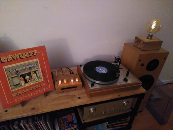 A low table with from left to right: upright the vinyl album Muscle Shoals by DeWolff, a Nixie clock, a spinning vinyl record on a player and a speaker.
Inside of the table, more vinyl records, some books and an amplifier can be seen.