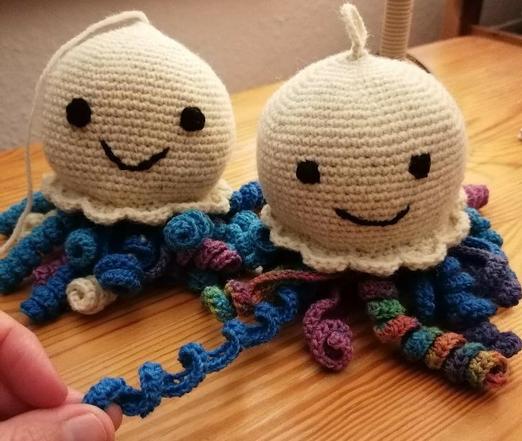 Two crocheted octopuses can be seen on a wooden desk surface. The head is crocheted in natural white sheep's wool with single crochet stitches. Two eyes and two smiling mouths are stitched on with black cotton yarn. The top of the head is worked in a kind of frill around the neck. The octopuses have lots and lots of curly legs in different colors. Different shades of blue, purple, natural white, multicolored. The smile of the left octopus seems to be bigger, but the right octopus also looks very friendly and happy. Each creature is unique :)
