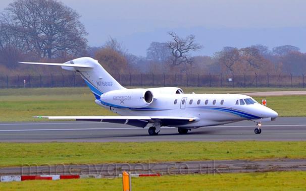 Side view of a white, 2 engined BizJet with the engines mounted on the sides of the rear fuselage, taxiing from left to right.
The plane has a grey, purple, and blue swoosh running from under the nose, along the body and the bottom of the engines, and up in to the tail.
There are silver "Citation X" titles on the engine pods, and the registration "N750GF" on the tail, also in silver.
Grass marks the edges of the runway and taxiways in the foreground, with more grass in the background leading up to trees in the distance, under grey skies.