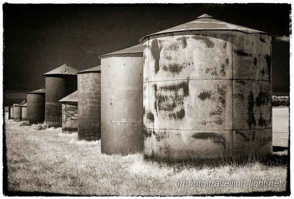 A toned black and white infrared photo of a row of feed silos of different sizes in a grassy field, seen from an oblique angle.