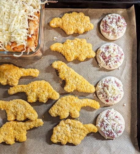 Blech mit Backpapier. Auf dem Blech liegen noch gefrorene, hellbraune Dino-Nuggets, Mini-Pizzen und eine gläserne Auflaufform mit Nudelauflauf und Streukäse.