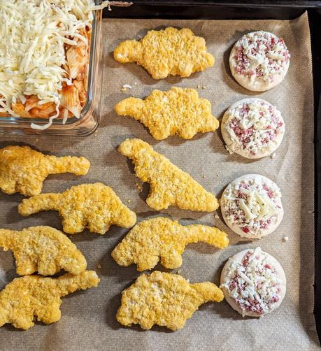 Blech mit Backpapier. Auf dem Blech liegen noch gefrorene, hellbraune Dino-Nuggets, Mini-Pizzen und eine gläserne Auflaufform mit Nudelauflauf und Streukäse.