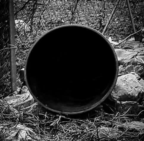 A black and white photo of the round opening of a trashcan which is laying on its side in an empty lot. The photo is taken from the open end of the can so as to appear to be a black circle. Grass, stones, broken concrete, and garbage litter the area surrounding the can.